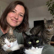 Eine Frau sitzt mit drei Katzen in ihrem Wohnzimmer. Eine schwarz-weiße Katze, eine getigerte Katze und eine weiß-getigerte Katze blicken gemeinsam in die Kamera.