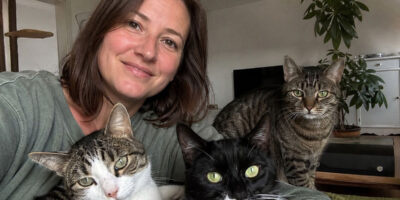 Eine Frau sitzt mit drei Katzen in ihrem Wohnzimmer. Eine schwarz-weiße Katze, eine getigerte Katze und eine weiß-getigerte Katze blicken gemeinsam in die Kamera.
