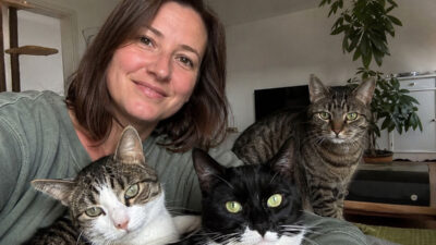 Eine Frau sitzt mit drei Katzen in ihrem Wohnzimmer. Eine schwarz-weiße Katze, eine getigerte Katze und eine weiß-getigerte Katze blicken gemeinsam in die Kamera.