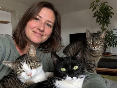 Eine Frau sitzt mit drei Katzen in ihrem Wohnzimmer. Eine schwarz-weiße Katze, eine getigerte Katze und eine weiß-getigerte Katze blicken gemeinsam in die Kamera.