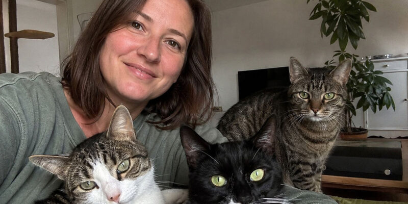 Eine Frau sitzt mit drei Katzen in ihrem Wohnzimmer. Eine schwarz-weiße Katze, eine getigerte Katze und eine weiß-getigerte Katze blicken gemeinsam in die Kamera.