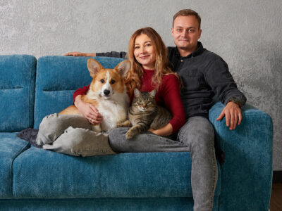 Ein Paar sitzt mit einem Corgi und einer getigerten Katze auf einem blauen Sofa und blickt in die Kamera.