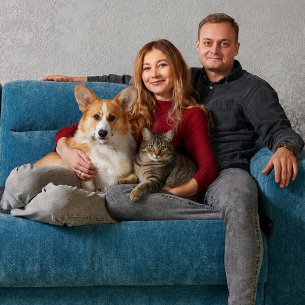 Ein Paar sitzt mit einem Corgi und einer getigerten Katze auf einem blauen Sofa und blickt in die Kamera.