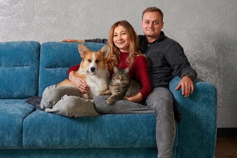 Ein Paar sitzt mit einem Corgi und einer getigerten Katze auf einem blauen Sofa und blickt in die Kamera.