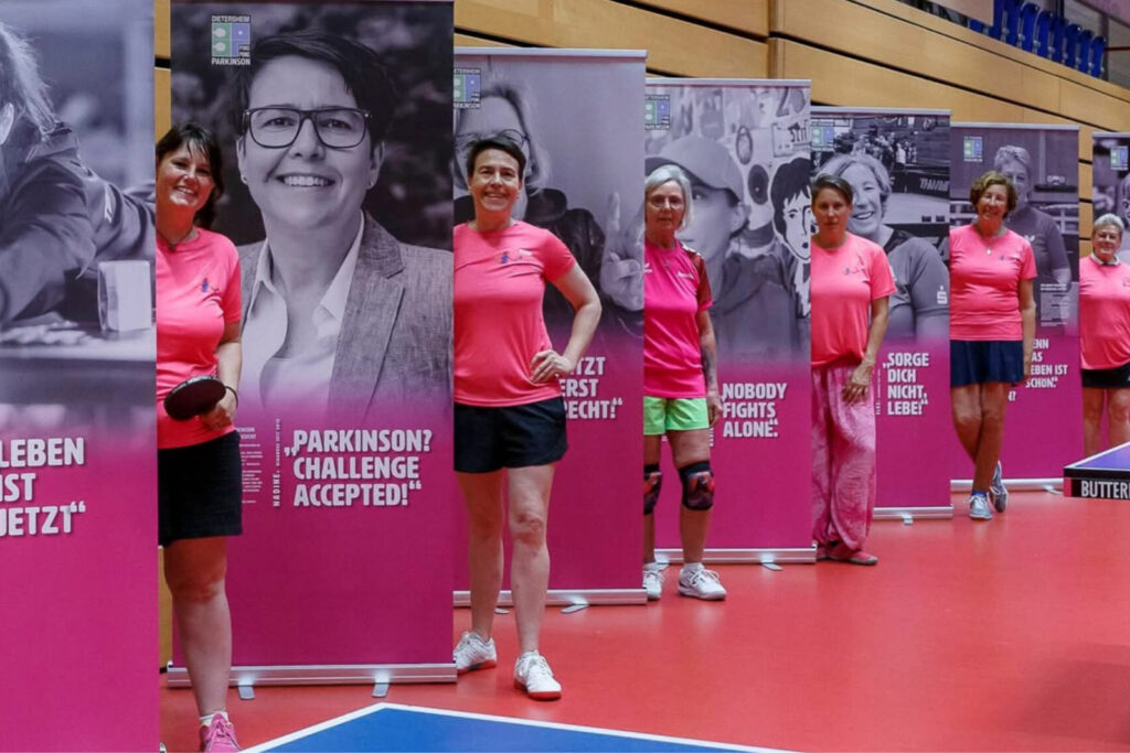 ping-pong-parkinson-frauen-gruppe Eine Gruppe von Frauen im Sportoutfit steht lächelnd an einer Tischtennisplatte in einer Turnhalle bei einem PingPongParkinson-Event.