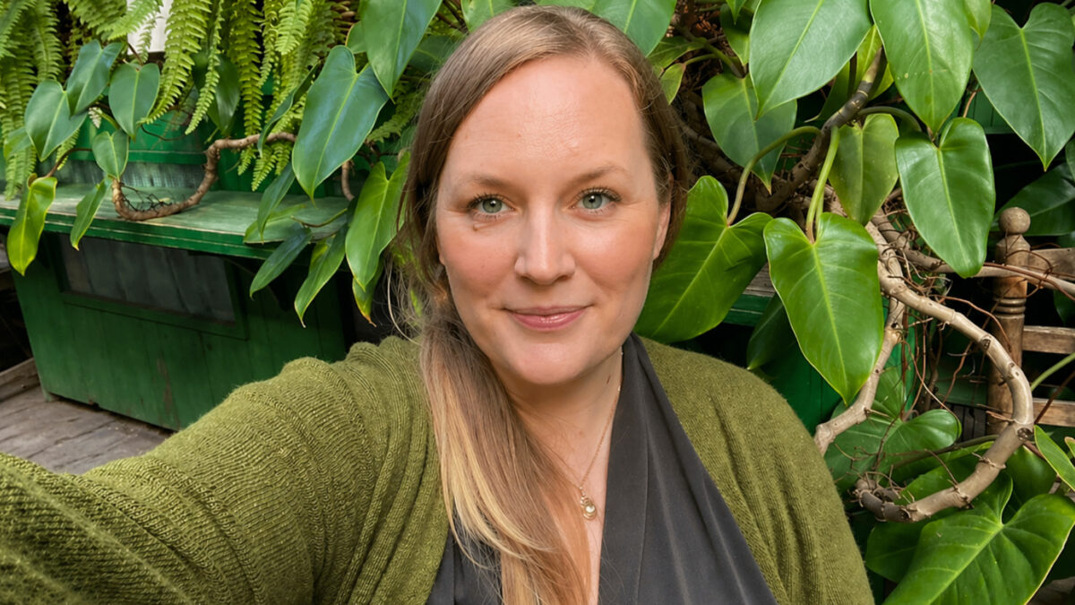 Porträt von Katharina, die lächelnd im Freien vor einer dichten Wand aus grünen Blättern posiert