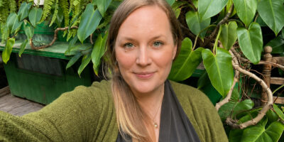 Porträt von Katharina, die lächelnd im Freien vor einer dichten Wand aus grünen Blättern posiert