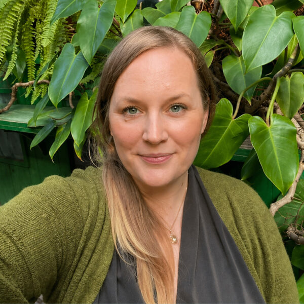 Porträt von Katharina, die lächelnd im Freien vor einer dichten Wand aus grünen Blättern posiert