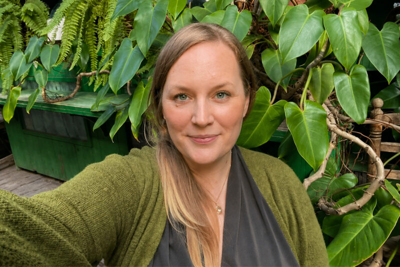 Porträt von Katharina, die lächelnd im Freien vor einer dichten Wand aus grünen Blättern posiert