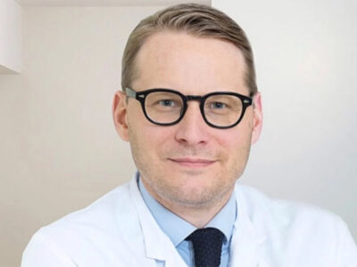 Porträt von Prof. Dr. Lars Wojtecki in Arztkleidung mit Brille vor hellem, neutralem Hintergrund.
