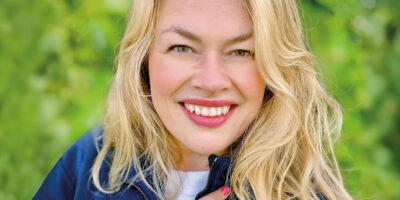 Porträt einer lächelnden blonden Frau (Nadine), die von chronischem Clusterkopfschmerz betroffen ist.