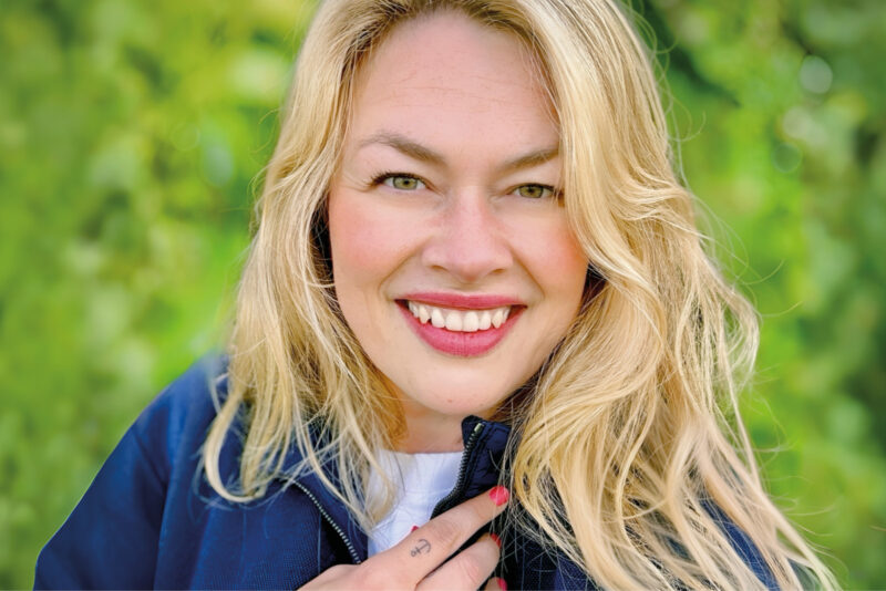 Porträt einer lächelnden blonden Frau (Nadine), die von chronischem Clusterkopfschmerz betroffen ist.