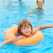 Kind mit orangefarbenem Schwimmring im Schwimmbad, im Hintergrund schwimmt eine erwachsene Person
