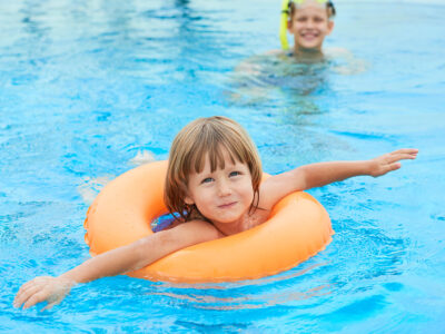 Kind mit orangefarbenem Schwimmring im Schwimmbad, im Hintergrund schwimmt eine erwachsene Person