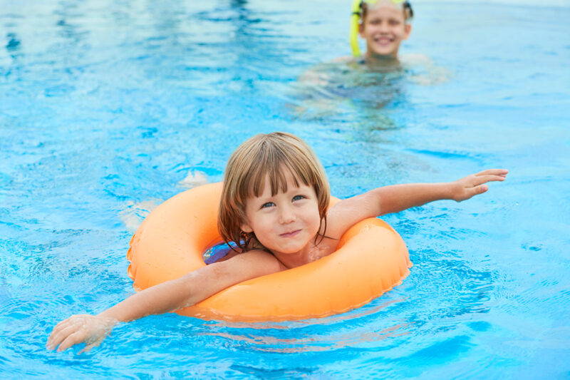 Kind mit orangefarbenem Schwimmring im Schwimmbad, im Hintergrund schwimmt eine erwachsene Person